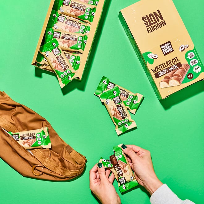 Hände öffnen Bio Schoko-Waffelriegel Crispy Hazel, daneben weitere Riegel und Boxen auf grünem Hintergrund