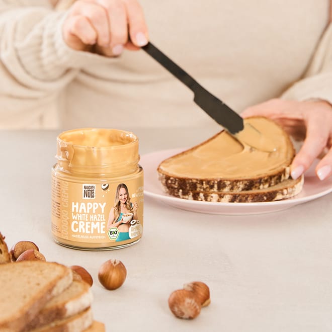 Ein Glas Happy White Hazel Creme und eine Hand, die die Creme auf ein Brot streicht