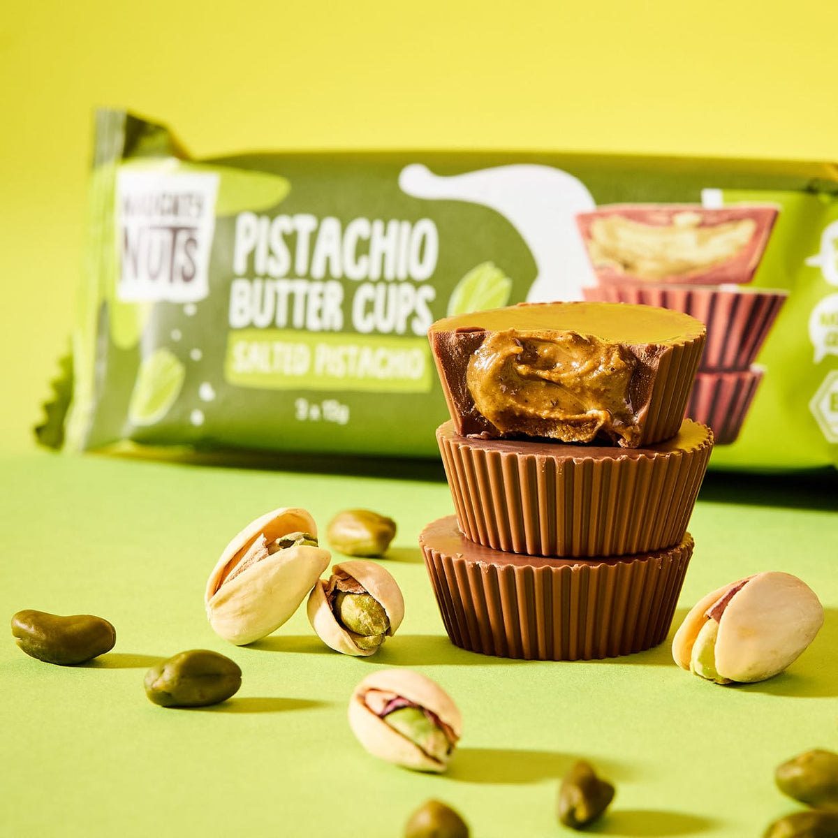 Drei gestapelte Bio-Nussbutter-Cups mit Pistazienfüllung und Pistazienkernen auf grünem Hintergrund