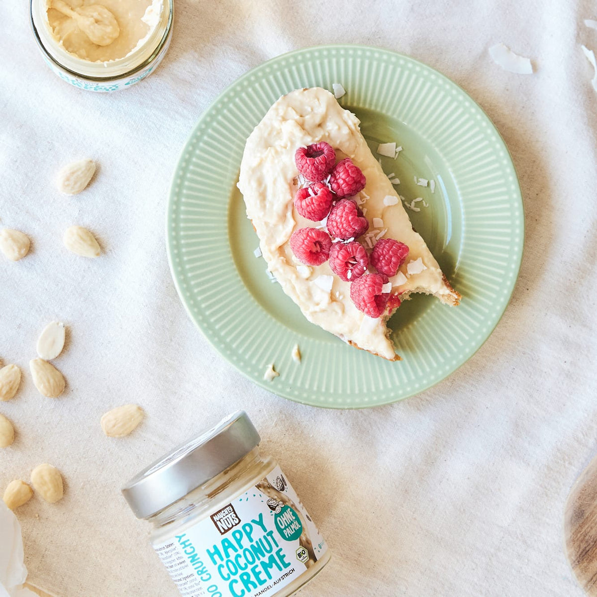 Happy Coconut Creme auf Brot mit Himbeeren und Kokosflocken, daneben ein Glas Happy Coconut Creme
