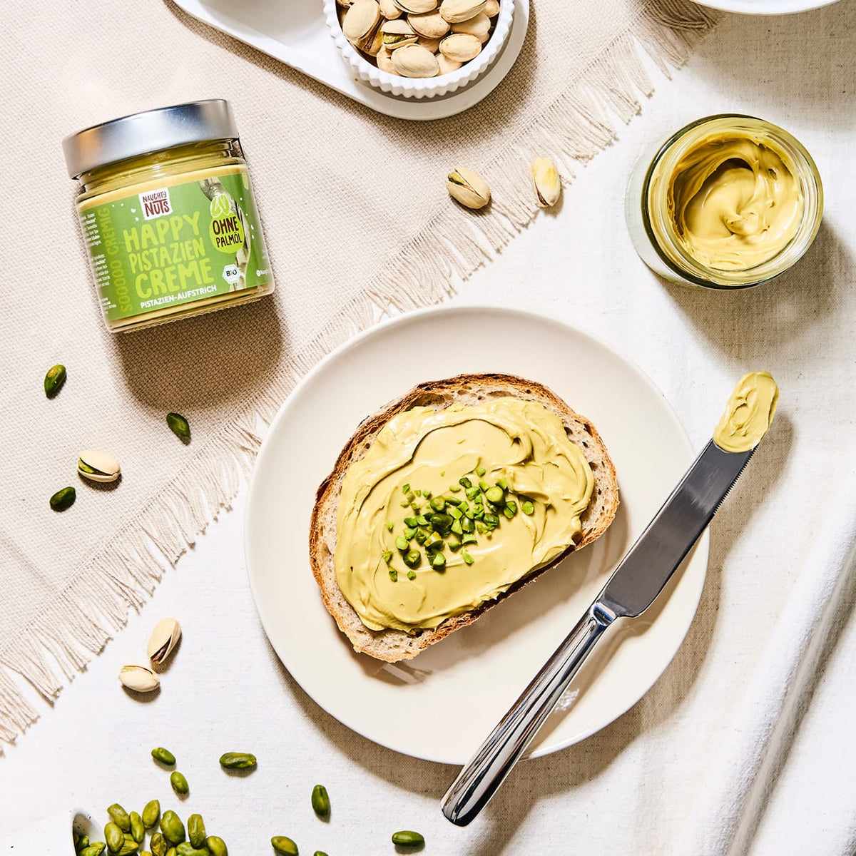 Happy Pistazien Creme auf Brot mit gehackten Pistazien, daneben ein Messer und ein Glas Creme